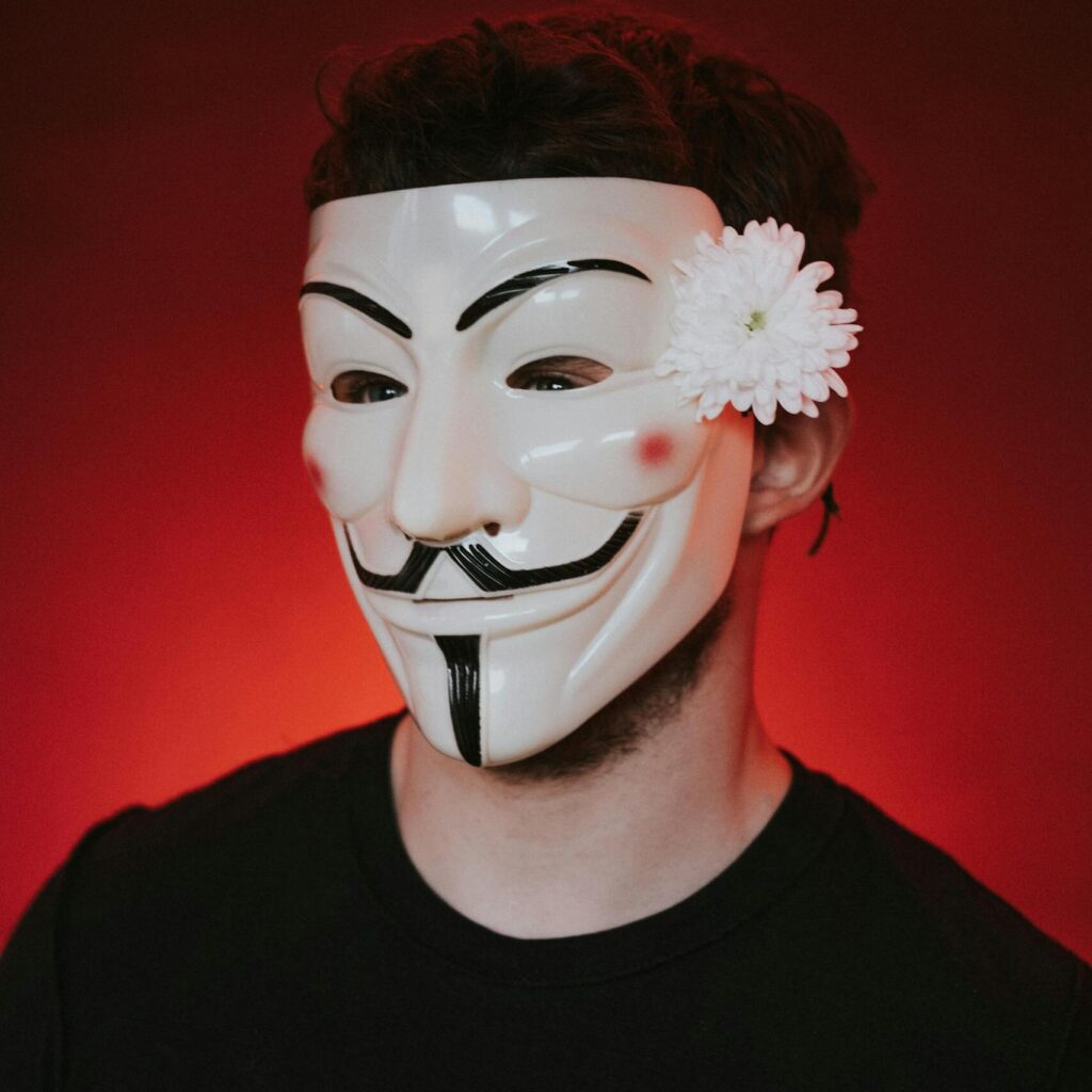 Male freedom fighter in black apparel and anonymous mask decorated with blooming white chrysanthemum looking forward